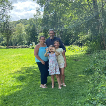 Eine lächelnde vierköpfige Familie steht gemeinsam auf dem grünen Rasen eines sonnigen Parks, umgeben von Bäumen. Das jüngste Mädchen winkt aufgeregt mit den Händen nach vorne.