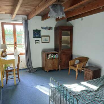 This cozy room features wooden beams, a window letting in sunlight, a bed, rustic wooden furniture, a wicker chair, books, and nautical-themed wall decor.