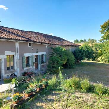 A charming countryside house with a tiled roof and green shutters sits amidst lush greenery, gardens, and trees under a clear blue sky. Peaceful ambiance.