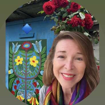 A smiling woman with brown hair and a colorful scarf stands in front of vibrant folk art, blooming red roses, and a rustic blue door outdoors.