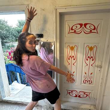 Une jeune femme pose de manière espiègle à l'intérieur, vêtue d'un t-shirt rose et d'un short noir. Elle se tient près d'une porte blanche ornée de motifs colorés rouges et jaunes, souriante.