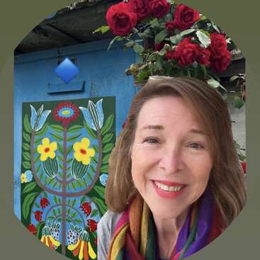 A smiling woman with brown hair and a colorful scarf stands in front of vibrant folk art, blooming red roses, and a rustic blue door outdoors.