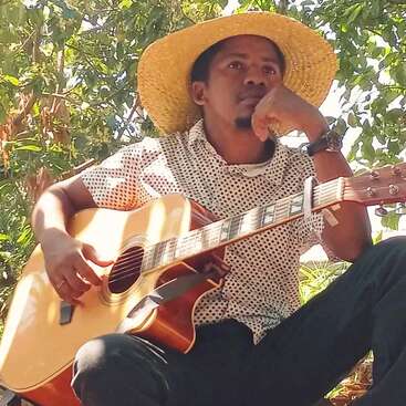 A man wearing a wide-brimmed straw hat and a polka dot shirt sits outdoors, thoughtfully holding an acoustic guitar, surrounded by green leafy trees and sunlight.