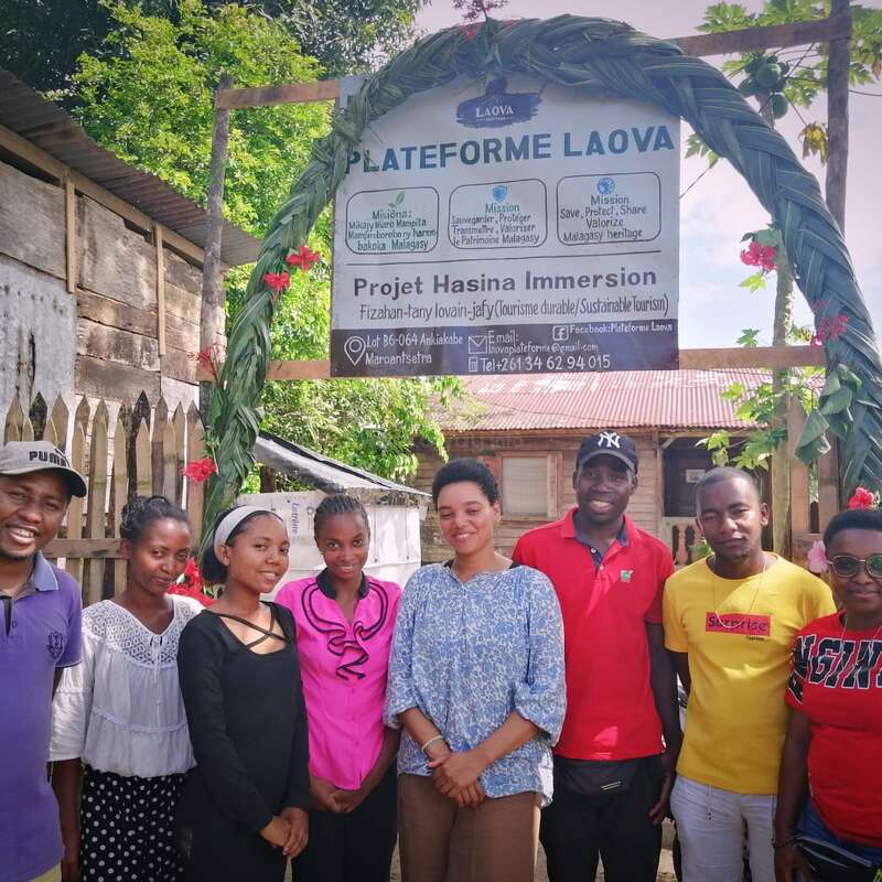 A group of eight people stands together smiling beneath a decorated arch with flowers, in front of a sign for Plateforme Laova and sustainable tourism.