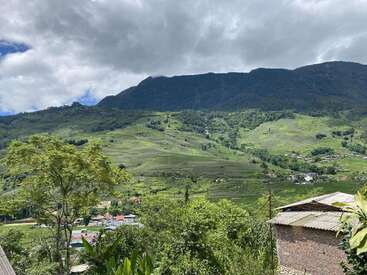 Ein saftig grünes Tal erstreckt sich unter dunklen, bewaldeten Bergen. Verstreute Häuser säumen die Landschaft. Dramatische Wolken schweben über dem Tal und werfen schummriges Sonnenlicht auf die lebendige Landschaft.