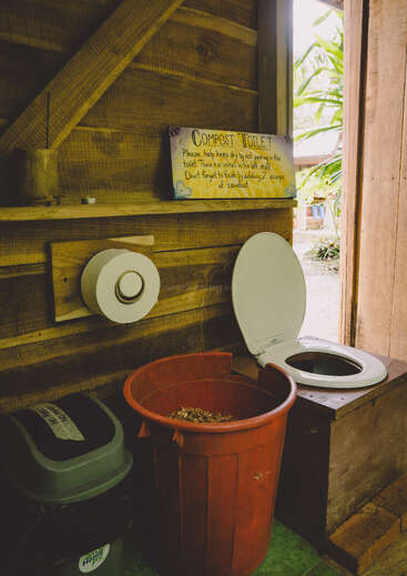 This image shows a rustic compost toilet inside a wooden structure, with a red sawdust bin, toilet paper, a recycling bin, and an instructional sign above.