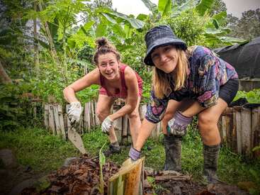 Two women happily gardening together, wearing gloves and boots, surrounded by lush green plants and trees, working with soil and compost in a vibrant backyard setting.