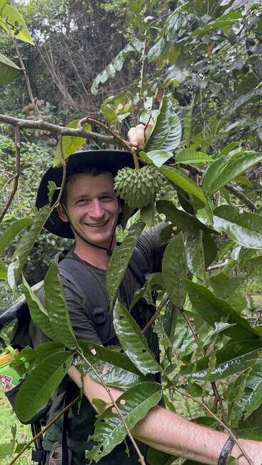 A smiling man in a black hat stands in dense jungle, holding a spiky green fruit among large, wet leaves. He wears a backpack and dark shirt.