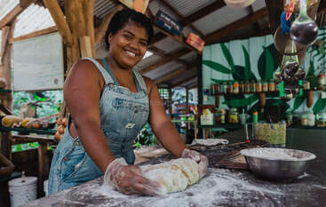 A woman in overalls smiles while kneading dough in a rustic, open kitchen. Colorful jars line the shelves, and sunlight filters through leafy surroundings outside.