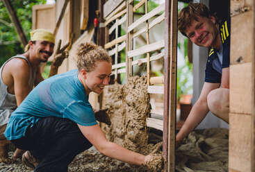 Three people smiling while working together on a construction project, applying a mixture to a wooden frame, possibly building an eco-friendly wall, outdoors in a natural setting.