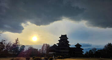Ein dramatischer Himmel mit dunklen Wolken und strahlendem Sonnenschein erstreckt sich über einer historischen japanischen Burg, die von Bäumen und Gärten umgeben ist, und schafft eine ruhige, majestätische Atmosphäre.