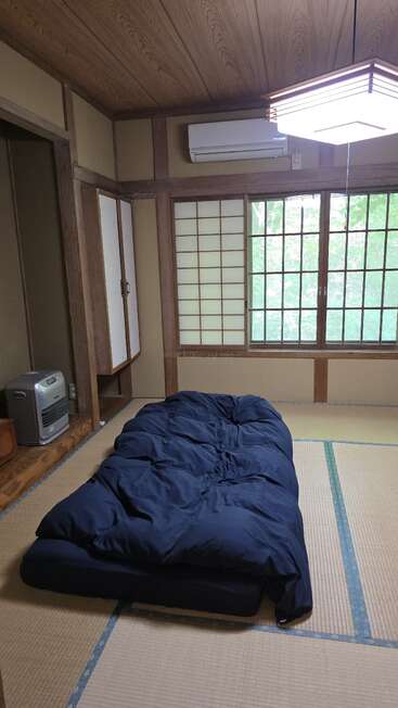 This image shows a traditional Japanese room with tatami mat flooring, a futon bed, sliding shoji doors, wooden ceiling, and a window with a nature view.