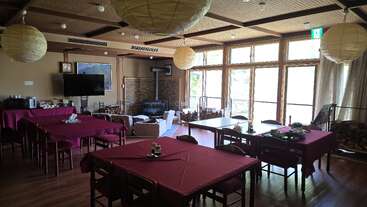 A cozy dining room with large windows, wooden floors, red tablecloths, paper lanterns, a fireplace, and comfortable seating, creating a warm, inviting atmosphere.