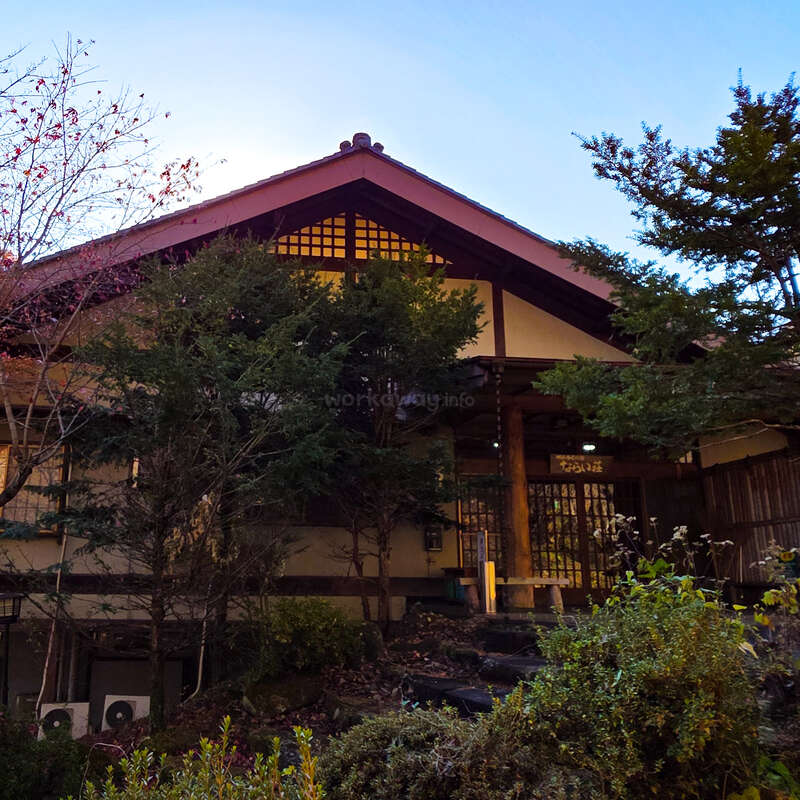 Ein traditionelles japanisches Haus, das teilweise von Bäumen und Sträuchern verdeckt wird, steht ruhig in der Abenddämmerung da. Das Holzhaus hat ein Satteldach und verzierte Gitterfenster.