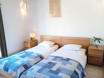 A imagem mostra um quarto sereno com duas camas de solteiro, cada uma com cabeceira de madeira e mesa de cabeceira, adornada com roupas de cama brancas e uma colcha azul, contra uma parede branca e nítida.