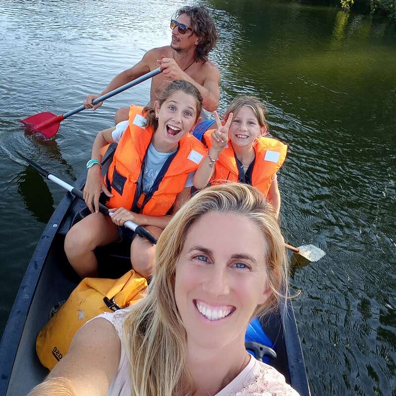 Uma família de quatro pessoas desfruta de um passeio de canoa em um lago tranquilo, com duas crianças usando coletes salva-vidas laranja e uma mulher tirando uma selfie em primeiro plano.