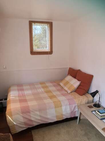 Um quarto aconchegante com um lençol xadrez rosa e amarelo, duas almofadas, uma mesinha lateral simples de madeira clara, um abajur preto, livros e uma janela pequena com uma persiana de bambu.