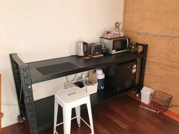 Uma cozinha compacta bem arrumada, com uma bancada de metal preta, micro-ondas, minigeladeira, torradeira, chaleira, banqueta branca, cestos para guardar coisas e vários utensílios de cozinha organizados com cuidado.