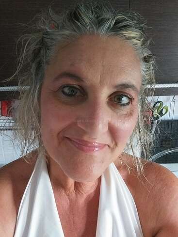 Une femme souriante aux yeux clairs et aux cheveux grisonnants et ondulés prend un selfie dans une cuisine. Elle porte un haut blanc et semble détendue et satisfaite.