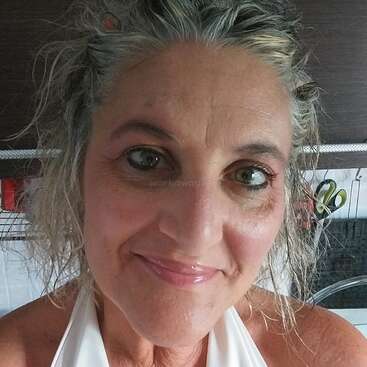 Eine lächelnde Frau mit hellen Augen und gewelltem, ergrauendem Haar macht ein Selfie in einer Küche. Sie trägt ein weißes Oberteil und wirkt entspannt und zufrieden.