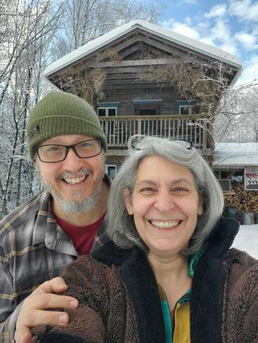 Um casal sorridente posa para uma selfie em frente a uma cabana rústica, cercada por árvores cobertas de neve e lenha. O clima é alegre, aconchegante e bem de inverno.