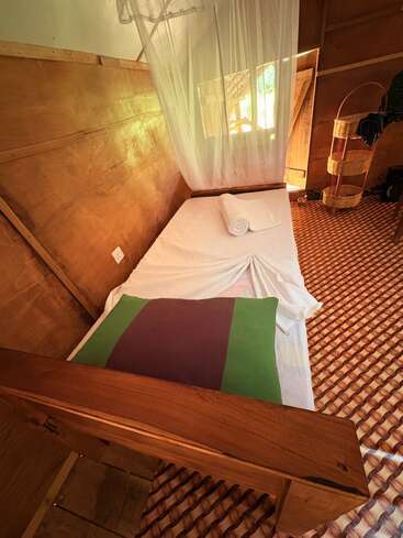 Cette image montre une chambre en bois simple et confortable avec un lit simple, une moustiquaire, une serviette enroulée, un sol à motifs et la lumière naturelle d'une fenêtre.