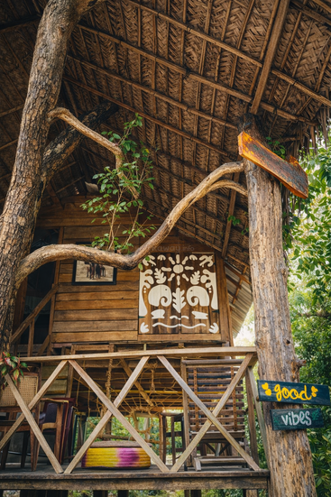 A rustic wooden treehouse with a thatched roof, surrounded by greenery. Artistic white mural, wooden furniture, rope swing, and a colorful "good vibes" sign create a relaxed atmosphere.