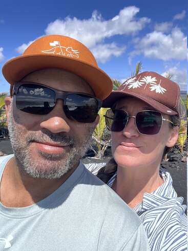 Una pareja sonriente posa para un selfie al aire libre. Ambos llevan gafas de sol y gorra. El cielo azul, las nubes esponjosas y las palmeras verdes sugieren un lugar tropical y soleado.