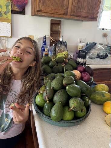 Una chica sonriente disfruta comiendo aguacate en una cocina, junto a un gran bol lleno de aguacates frescos, con varias frutas y artículos de cocina de fondo.