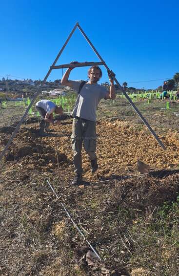 Un homme se tient debout dans un champ, tenant un grand cadre triangulaire, tandis qu'une autre personne travaille à proximité. On aperçoit un ciel bleu éclatant, de jeunes plants et des tuyaux verts.