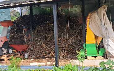 Une personne travaille près d'un grand tas de branches et de bûches, sous une structure couverte. On aperçoit des machines et des cultures, ce qui laisse supposer un environnement agricole en plein air.
