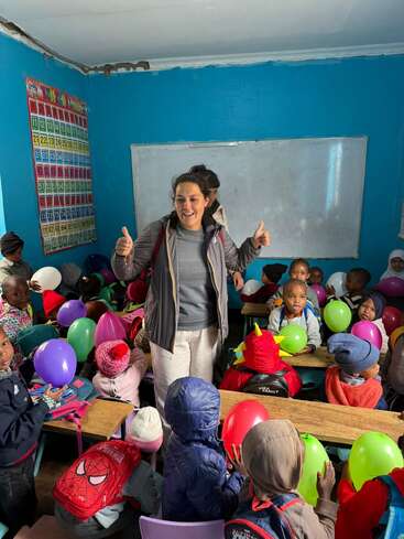 Uma mulher está sorrindo, levantando o polegar, cercada por crianças pequenas segurando balões coloridos em uma sala de aula alegre com paredes azuis e pôsteres educativos.