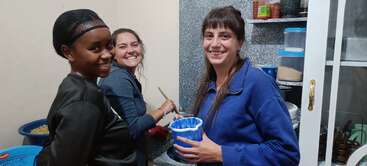 Três mulheres estão sorrindo e juntas em uma cozinha. Uma delas está lavando pratos, enquanto as outras ajudam e seguram recipientes. A atmosfera é amigável e alegre.