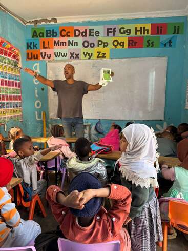 Um professor instrui energicamente jovens alunos em uma sala de aula colorida, decorada com o alfabeto e os números, enquanto as crianças ouvem atentamente e participam da aula.