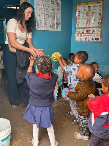 A woman interacts warmly with a group of young children in a colorful classroom, handing out balls while educational posters decorate the blue walls behind them.