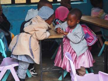Two young girls interact at a classroom table. One leans forward, curious, while the other, dressed in pink, concentrates on something in her hands. Others observe.