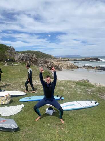 Un homme en combinaison de plongée prend une pose espiègle, la langue tirée, près de planches de surf posées sur l'herbe, au bord d'une plage rocheuse, sous un ciel partiellement nuageux.