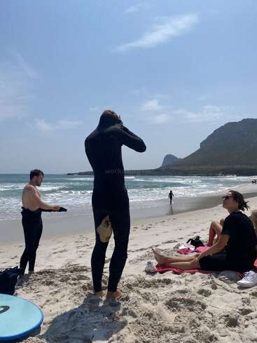 Deux personnes en combinaison de plongée se tiennent debout sur une plage de sable ; l'une d'elles ajuste sa cagoule. Deux autres sont assises sur une serviette, en train de se détendre. On aperçoit les vagues de l'océan et les montagnes en arrière-plan.