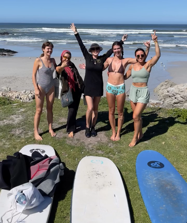 Cinq femmes sourient et prennent la pose, rayonnantes de joie, devant la plage, leurs planches de surf posées sur l'herbe, prêtes à passer une journée ensoleillée à surfer et à s'amuser.