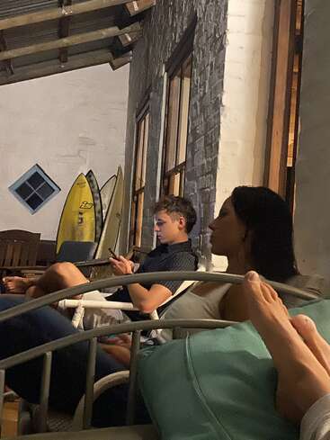 Two people relax on a porch, one using a phone, the other leaning back. Surfboards are propped against the wall, giving a laid-back, beach vibe.