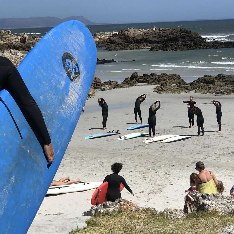 Un groupe de surfeurs en combinaison s'étire sur une plage de sable, près de leurs planches de surf, avec en arrière-plan des falaises rocheuses, les vagues bleues de l'océan et une grande planche bleue au premier plan.