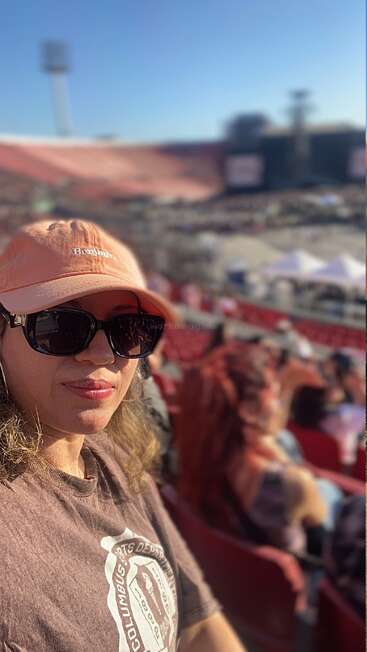 Uma mulher usando um boné pêssego e óculos escuros tira uma selfie em um estádio. As arquibancadas e o palco ao fundo sugerem um show.