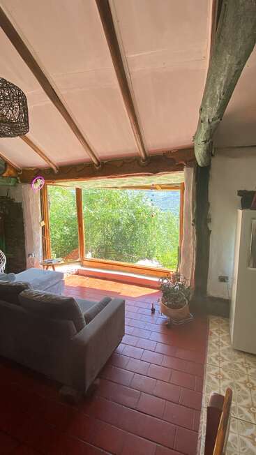 Sala de estar aconchegante com vigas rústicas, piso de ladrilho vermelho, sofá cinza, janela grande com vista para o verde exuberante do exterior, luz solar abundante, vaso de plantas e decoração natural e terrosa.