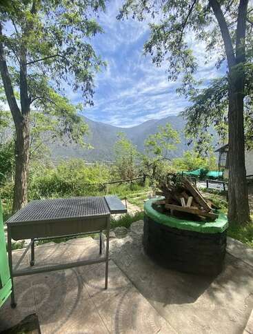Uma cena tranquila ao ar livre com uma churrasqueira, uma fogueira circular cheia de madeira, cercada por árvores verdes, com montanhas e céu azul ao fundo.