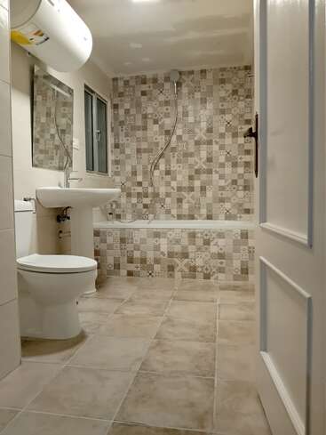 Dieses Badezimmer hat beige gemusterte Fliesen, eine Badewanne mit Dusche, eine weiße Toilette, ein Waschbecken, einen Spiegel, einen Warmwasserbereiter, ein Fenster und eine teilweise offene weiße Tür. Sauber und modern.