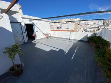 Dieses Bild zeigt eine sonnige Dachterrasse mit Topfpflanzen, einem kleinen roten Tisch und offenen Türen. Im Hintergrund sind weiße Gebäude und der blaue Himmel zu sehen.