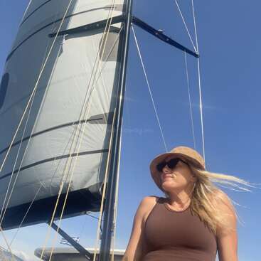 Eine Frau mit Sonnenbrille und Sonnenhut genießt einen sonnigen Tag auf einem Segelboot. Das große Segel und der klare blaue Himmel machen die entspannte, windige Szene komplett.