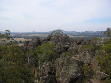 Das Bild zeigt einen zerklüfteten Felsen, umgeben von üppigem Grün, vor einer malerischen Landschaft mit sanften Hügeln und Feldern unter einem bewölkten Himmel.