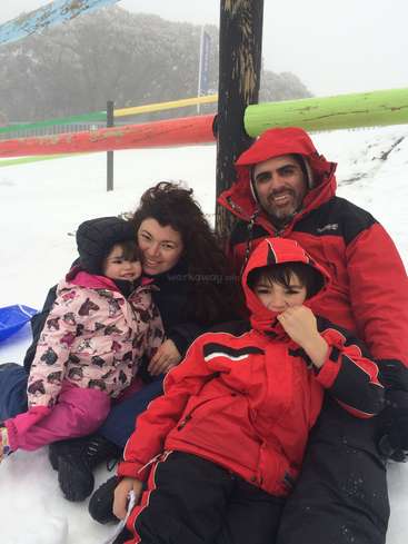Das Bild zeigt eine vierköpfige Familie, die auf dem schneebedeckten Boden sitzt, umgeben von einem bunten Zaun und einer Bergkulisse, mit einer ruhigen und friedlichen Atmosphäre.