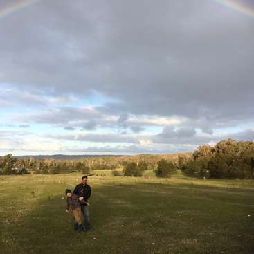 Das Bild zeigt eine heitere Szene mit einem Mann und einem Kind, die in einem üppigen grünen Feld stehen, während sich ein Regenbogen über ihnen spannt.